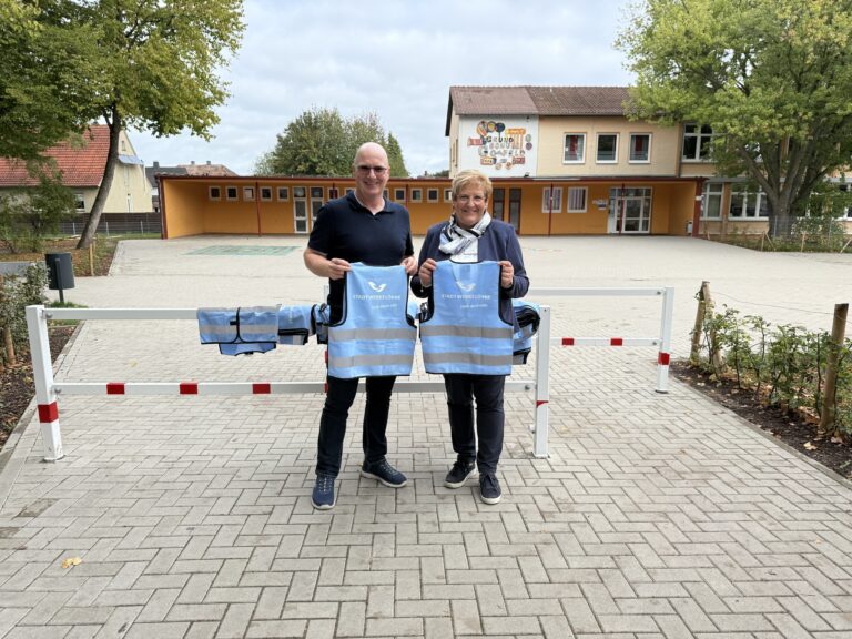 Der Betriebsleiter und die Rektorin stehen vor der Grundschule und halten zwei Warnwesten für Kinder.