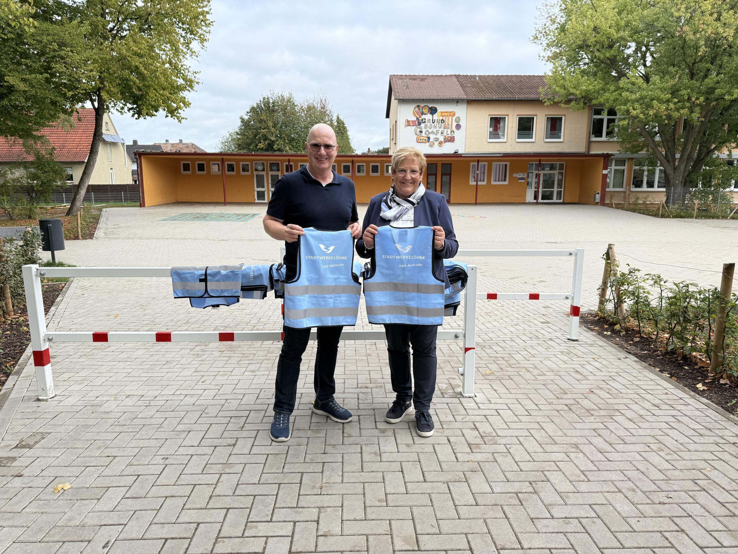 Der Betriebsleiter und die Rektorin stehen vor der Grundschule und halten zwei Warnwesten für Kinder.